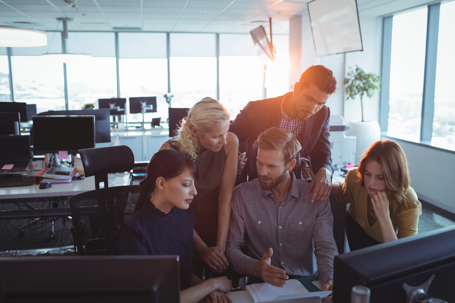 Five business colleagues gathered around a desk in a bright open-plan office, looking intently at a computer screen during a collaborative discussion.