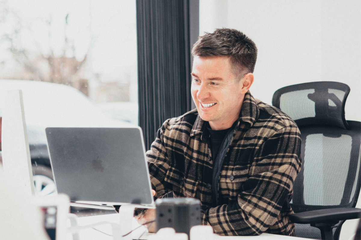 A smiling professional sits at a desk in a bright office, looking at a laptop screen. They are wearing a plaid overshirt and seated in a mesh-backed office chair near a large window. The workspace appears modern, clean, and well-lit with natural light.