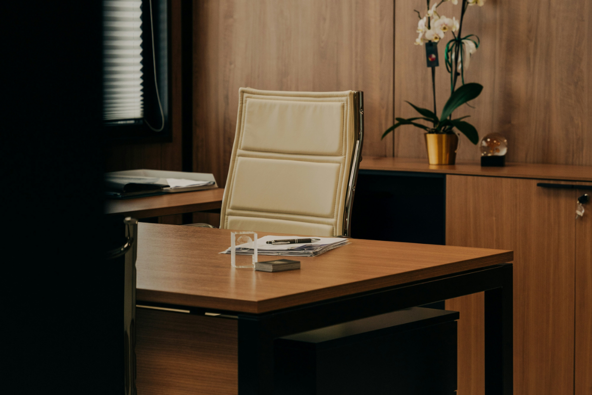 A modern executive office with a wooden desk and a cream-colored leather office chair. Papers and a pen rest neatly on the desk, while a potted orchid and decorative object sit on a wooden cabinet in the background. The space is tidy, professional, and warmly lit.