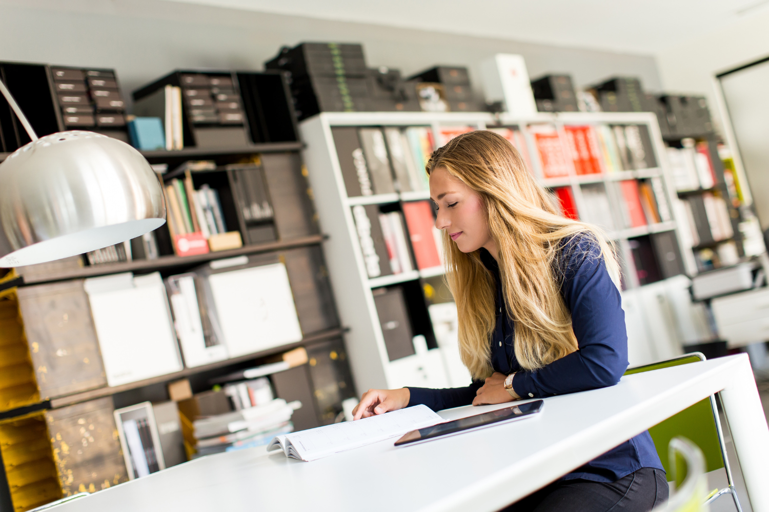 A woman with long blonde hair in a navy shirt leaning over a white desk, writing in a planner next to a tablet, with shelves of files and boxes behind her.