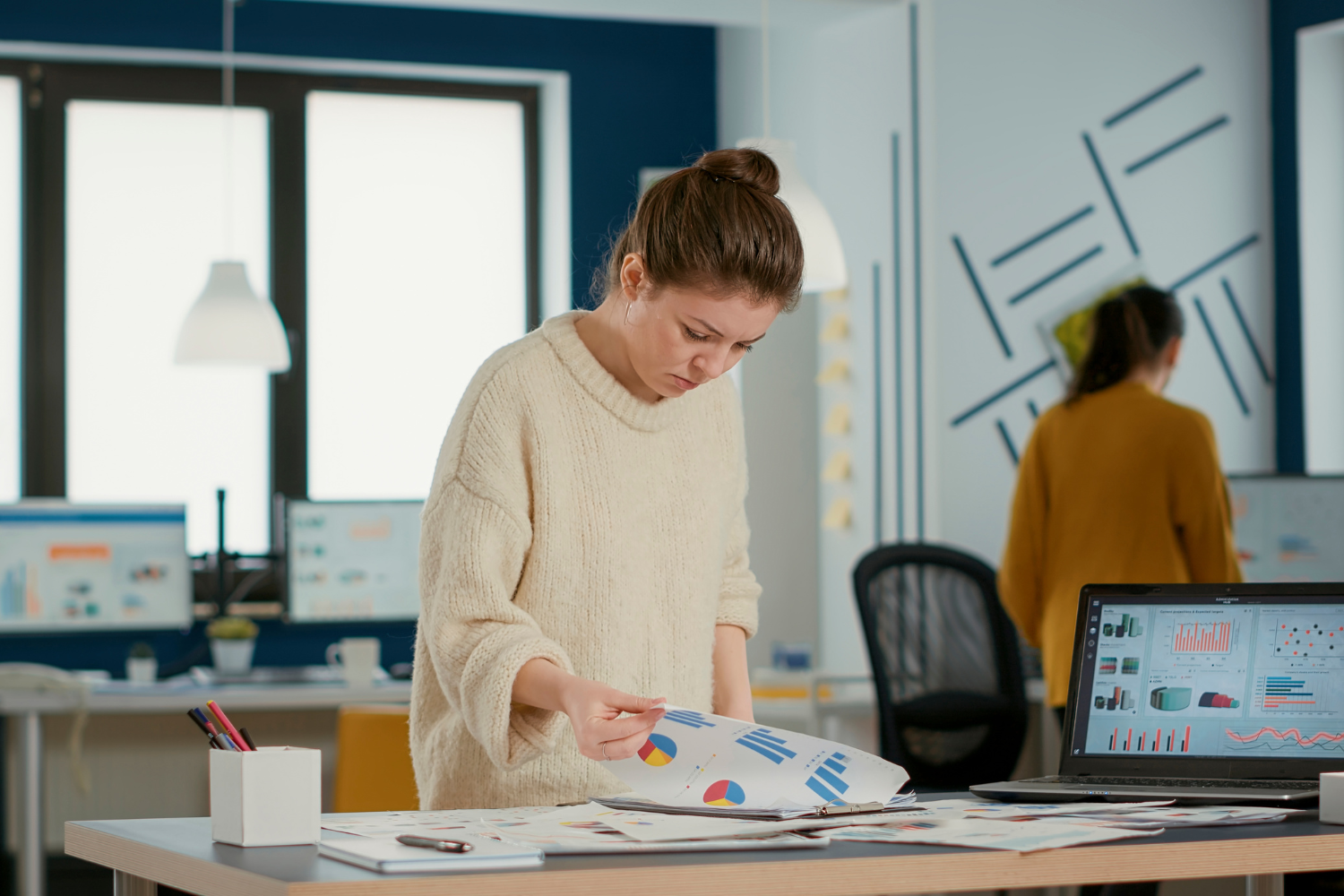 A woman in a cream sweater standing at a desk reviewing printed charts and graphs, with a laptop showing colourful data dashboards beside her in a modern office.