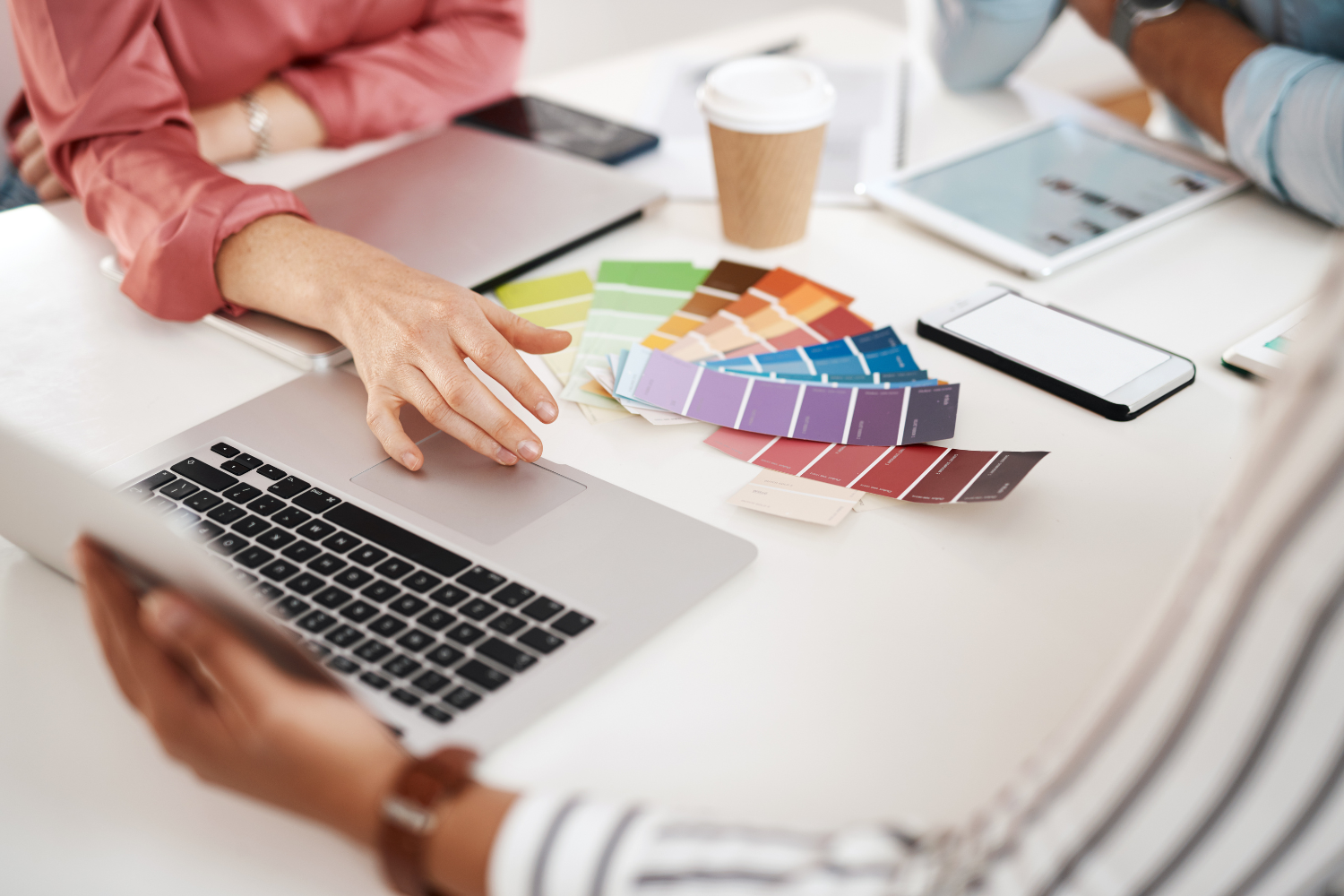 Close-up of two designers at a white table, one using a laptop trackpad beside a fanned-out set of colour swatch samples, a coffee cup, and a tablet.