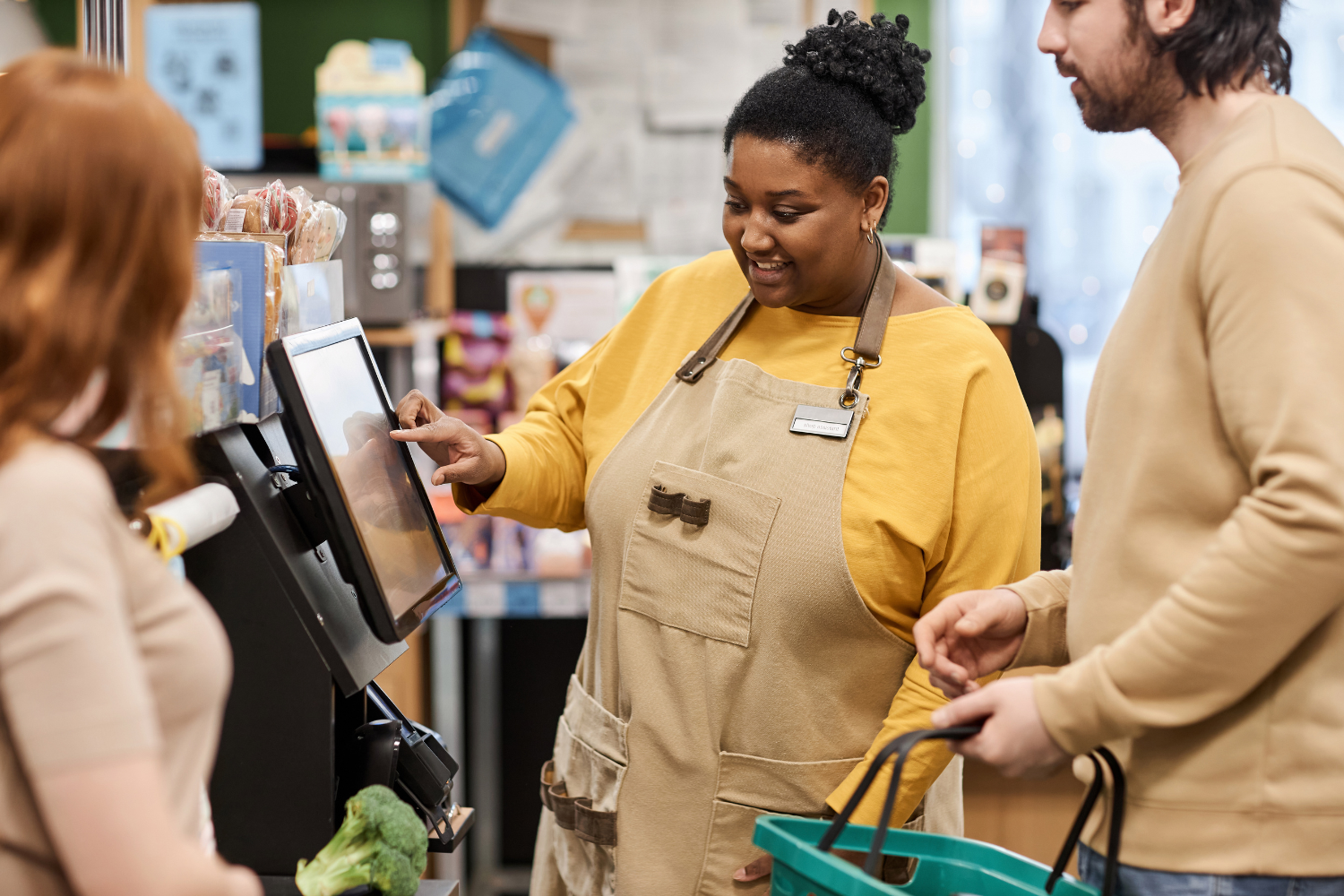 A smiling shop assistant in a yellow top and beige apron operating a touchscreen checkout to help two customers, one holding a green shopping basket, in a grocery store.