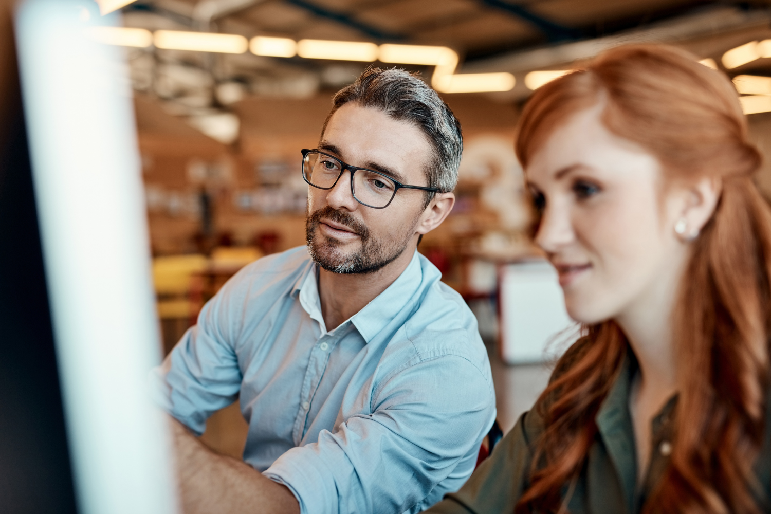 A man in glasses and a light blue shirt and a woman with red hair looking attentively at a screen together in a warmly lit workspace.