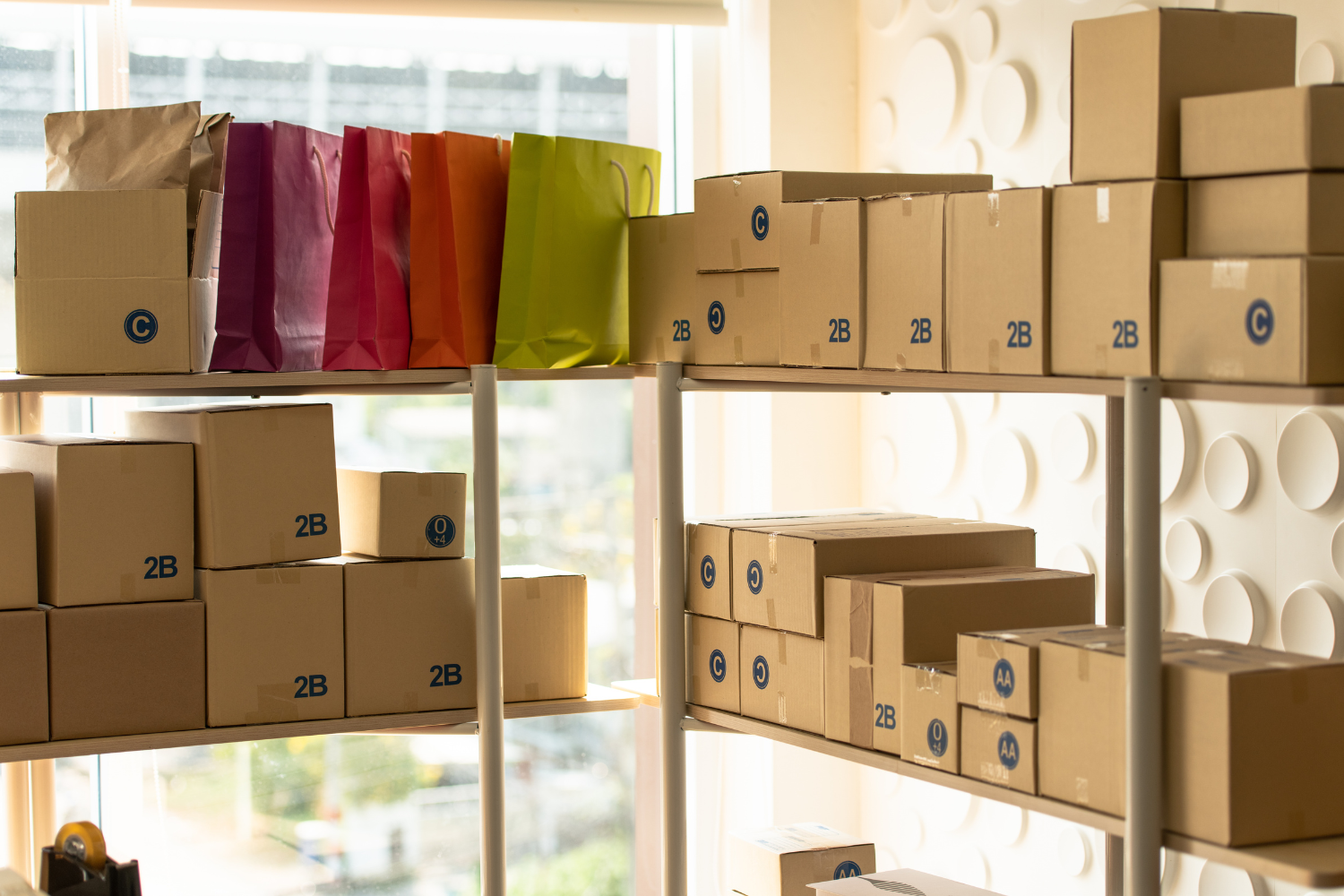 Metal shelving stacked with cardboard parcels labelled "2B" and other codes, alongside brightly coloured paper gift bags, beside a window.