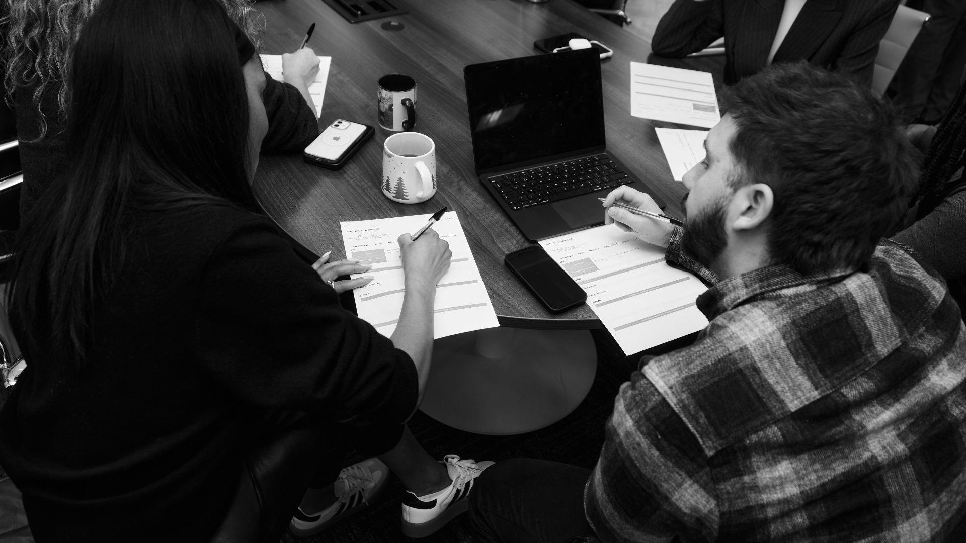 Black and white overhead photo of a business meeting: several people seated around a wooden table filling out worksheets by hand, with a laptop, two mugs, and two smartphones on the table between them.