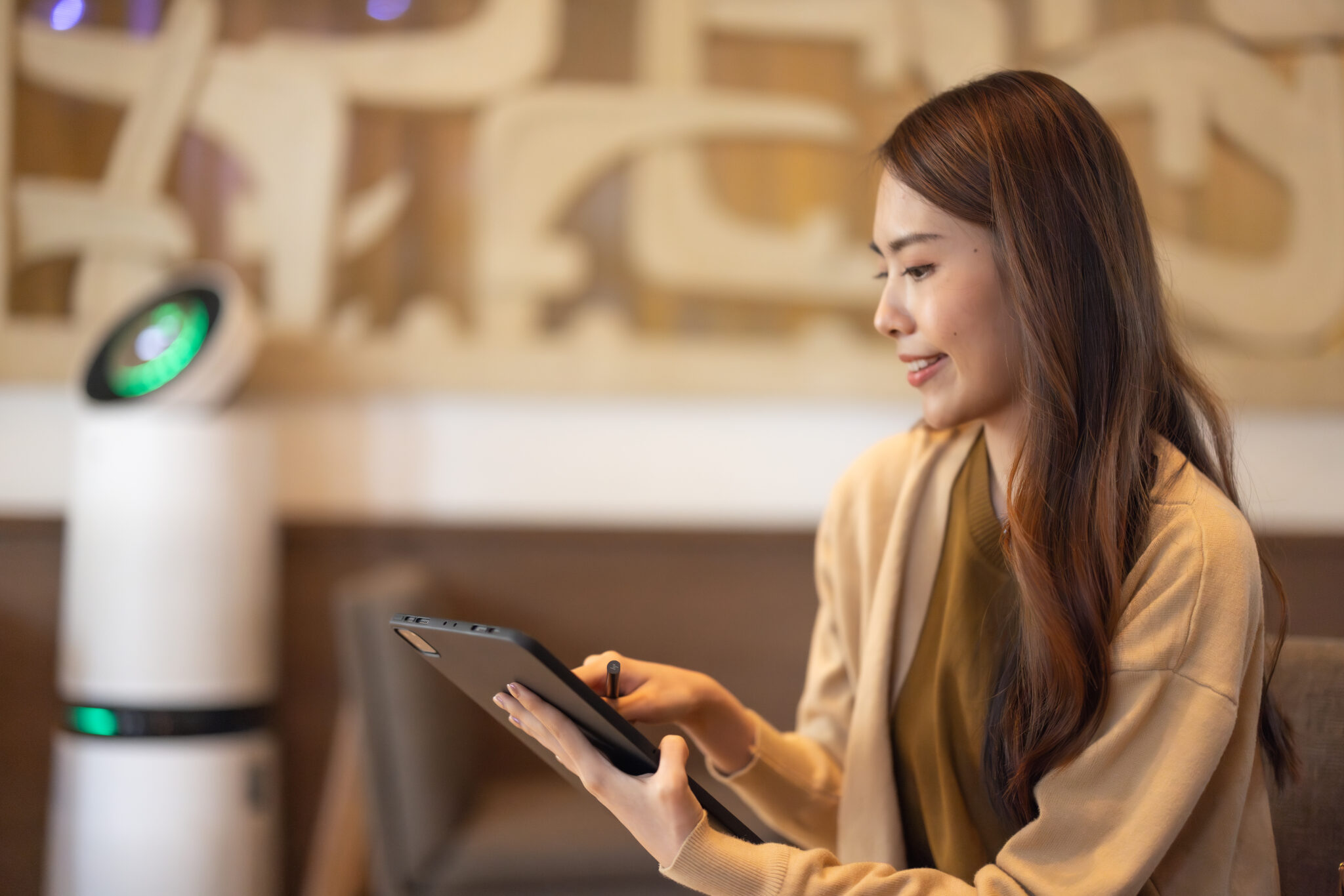 A young woman with long, brown hair smiles as she looks at a tablet in her hands. The setting appears to be indoors, possibly in a restaurant or workplace. In the blurred background, there is a white robot with a green screen. The photo has a warm, inviting tone and the woman appears to be focused and content. This picture could be used in projects about technology, robotics, or the future of work. Also could work well in project about lifestyle and young adults.